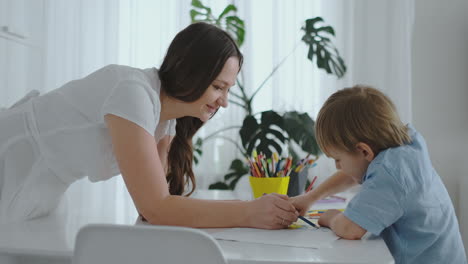 Young-beautiful-mother-and-son-draw-with-colored-pencils-sitting-at-the-table-in-the-kitchen.-The-camera-moves-in-slow-motion